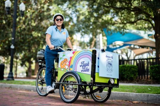 A female library staff member riding a three wheeled cargo bicycle, the cargo box is covered in colorful graphics and the label "Book Bike"