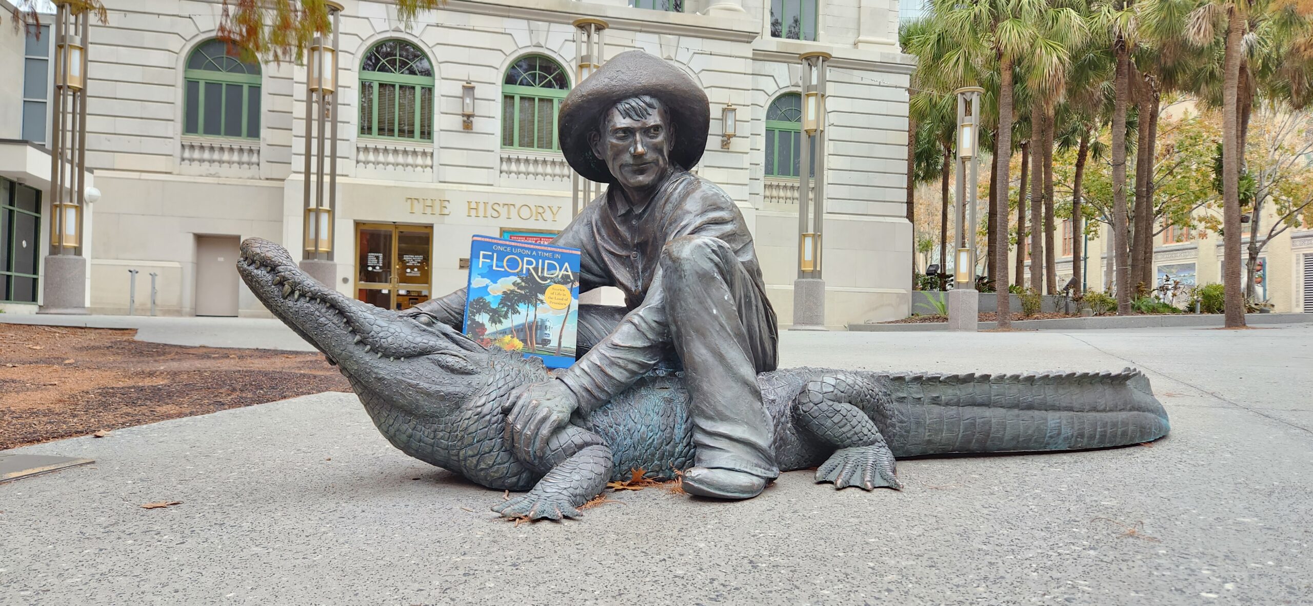 The book "Once Upon a Time in Florida" on top of a bronze statue of a man sitting on an alligator, located in Heritage Square in front of the Orange County Regional History Center.