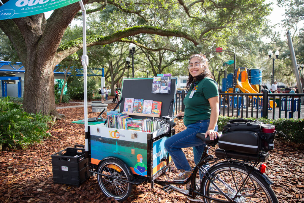 Outreach Coordinator, Nathalie Ruiz, sitting on the OCLS book bike in a park.